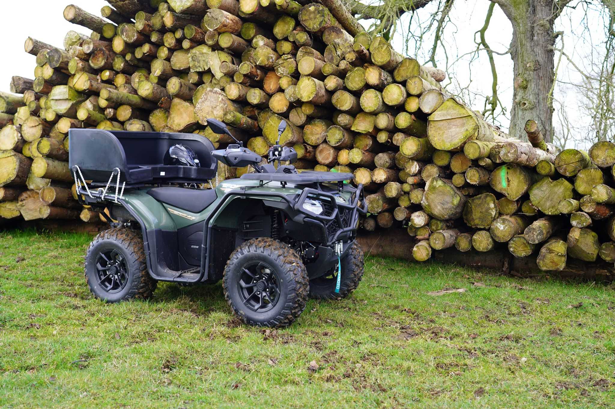 CFMOTO ATV quad bike with rear cargo box parked next to stacked timber logs in forestry setting